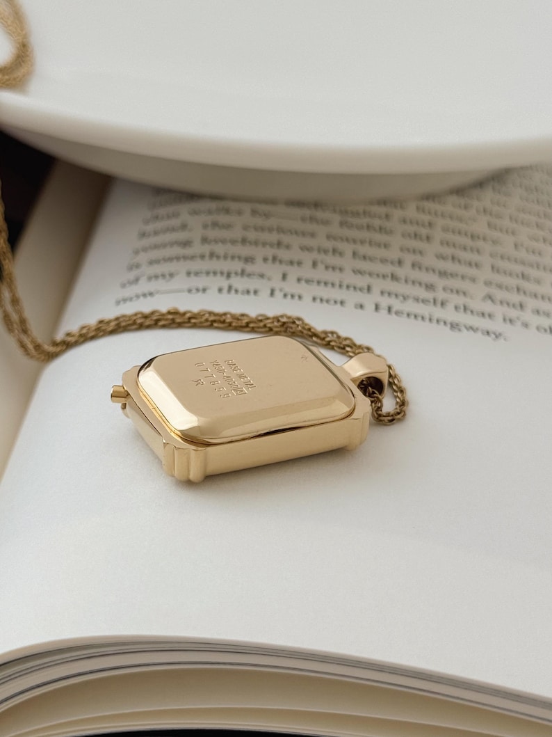 Puede incluir: Un collar con colgante rectangular dorado con una cadena delicada. El colgante tiene un dise&ntilde;o con bisagras y parece estar grabado con texto. El collar descansa sobre las p&aacute;ginas abiertas de un libro.