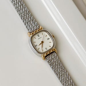 May include: A Seiko wristwatch featuring a square white dial with black Roman numeral indices and gold-tone hands. The watch case is silver-tone with gold-tone accents at the lugs, attached to a silver-tone woven metal mesh strap.