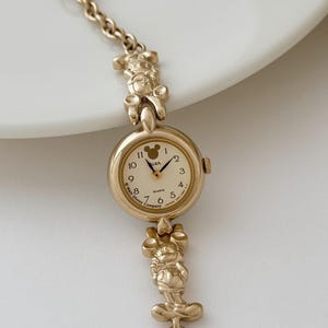 Puede incluir: Reloj de cuarzo ALBA dorado con diseño de Mickey Mouse. El reloj tiene una esfera redonda con números arábigos y una silueta de Mickey Mouse. La correa incluye figuras de Mickey Mouse. Un objeto de colección de Disney.