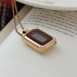 Puede incluir: Un colgante de reloj ALBA rectangular dorado con una esfera burdeos, suspendido de una delicada cadena dorada. El reloj muestra la hora digital. El colgante descansa sobre un libro abierto con el t&iacute;tulo "HEMINGWAY".
