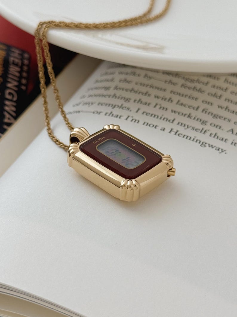 Puede incluir: Un colgante de reloj ALBA rectangular dorado con una esfera burdeos, suspendido de una delicada cadena dorada. El reloj muestra la hora digital. El colgante descansa sobre un libro abierto con el t&iacute;tulo "HEMINGWAY".