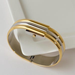 Puede incluir: Un reloj de pulsera rígida con bisagra que presenta una banda exterior dorada pulida y una banda interior plateada. La esfera del reloj está integrada en la parte superior de la pulsera, mostrando un pequeño dial rectangular con una corona dorada en el lateral.