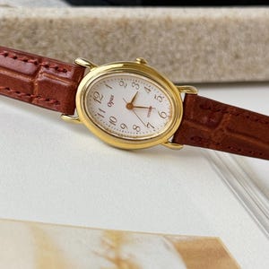 May include: An oval-shaped gold-tone wristwatch with a white dial featuring black Arabic numerals, gold hands, and the text Opus and Citizen. The watch has a brown leather strap with a textured finish and visible stitching along the edges.