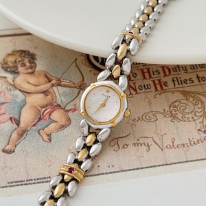 May include: A vintage Tisse watch with a white face and gold-tone accents. The watch band features a mix of silver and gold-tone links. A small red jewel is set in the band near the watch face. The watch is displayed on a vintage Valentine's Day card.
