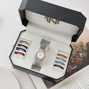 Op de afbeelding: Een zilverkleurig Anne Klein horloge met een witte wijzerplaat en een mesh band, gepresenteerd in een zwart-witte doos. De doos bevat een selectie van verwisselbare gekleurde ringen. Het horloge wordt getoond op een open boek.