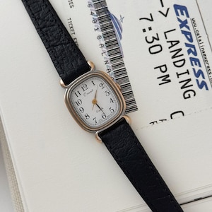 May include: A vintage Orient quartz watch with a rectangular gold-toned case and a white dial featuring black numerals. The watch has a black leather strap. The watch is displayed on a white surface with a Catalina Express ticket.