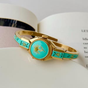 Puede incluir: Un reloj tipo brazalete dorado con una esfera redonda sobre un fondo de esmalte turquesa brillante y un motivo central de flor de edelweiss. La banda articulada incluye segmentos de esmalte turquesa a juego decorados con pequeños motivos florales verdes.