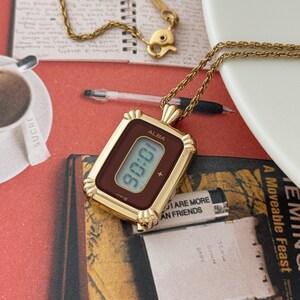 Puede incluir: Un collar con colgante de reloj dorado, de forma rectangular, que muestra la hora 90:06. El reloj tiene una esfera marr&oacute;n y est&aacute; suspendido de una delicada cadena dorada. El reloj tiene la marca "ALBA".