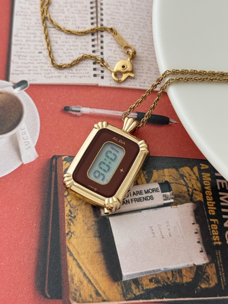 Puede incluir: Un collar con colgante de reloj dorado, de forma rectangular, que muestra la hora 90:06. El reloj tiene una esfera marr&oacute;n y est&aacute; suspendido de una delicada cadena dorada. El reloj tiene la marca "ALBA".
