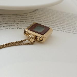 Puede incluir: Un collar con colgante dorado con un dije cuadrado que se asemeja a un reloj digital vintage. El dije tiene una esfera marr&oacute;n oscuro y una peque&ntilde;a pantalla te&ntilde;ida de verde. La cadena es delicada y tambi&eacute;n dorada.