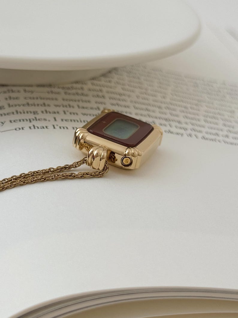 Puede incluir: Un collar con colgante dorado con un dije cuadrado que se asemeja a un reloj digital vintage. El dije tiene una esfera marr&oacute;n oscuro y una peque&ntilde;a pantalla te&ntilde;ida de verde. La cadena es delicada y tambi&eacute;n dorada.