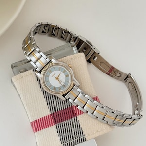 Könnte beinhalten: Eine silber- und goldfarbene Seiko-Armbanduhr mit hellblauem Zifferblatt und römischen Ziffern. Die Uhr wird auf einem kleinen, karierten Stoffkissen präsentiert. Das Armband ist silberfarben mit goldfarbenen Akzenten.