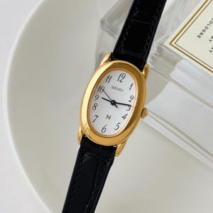 May include: A Seiko wristwatch with an oval gold-tone case and a white dial featuring black Arabic numerals. The watch has black hands and a black leather strap with a textured finish. The brand name Seiko is printed on the dial in black text.