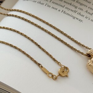 Puede incluir: Un collar dorado con una cadena y un cierre delicados, apoyado en un libro. El collar presenta un peque&ntilde;o colgante. El fondo incluye texto del libro.