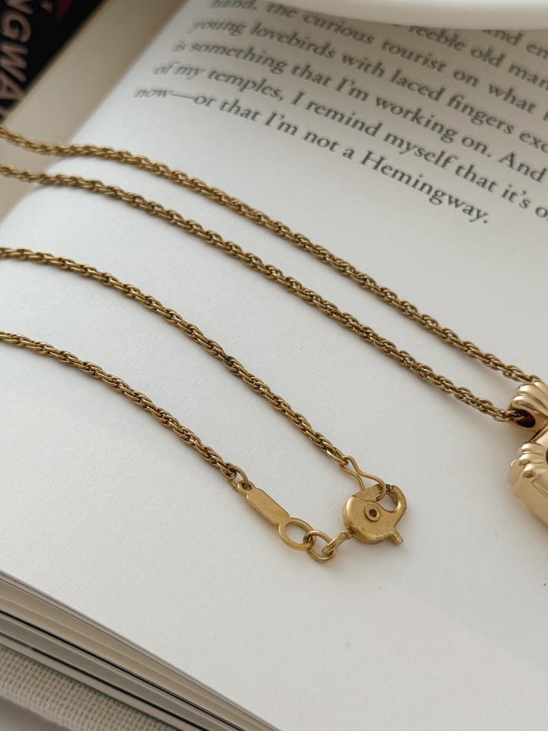 Puede incluir: Un collar dorado con una cadena y un cierre delicados, apoyado en un libro. El collar presenta un peque&ntilde;o colgante. El fondo incluye texto del libro.