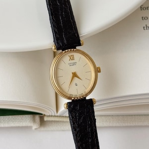 Könnte beinhalten: Eine goldfarbene Citizen Quartz Armbanduhr mit rundem Gehäuse, strukturiertem cremefarbenem Zifferblatt, goldenen Zeigern und einer römischen Ziffer auf der Zwölf-Uhr-Position. Die Uhr ist an einem schwarzen Lederarmband mit Struktur befestigt.