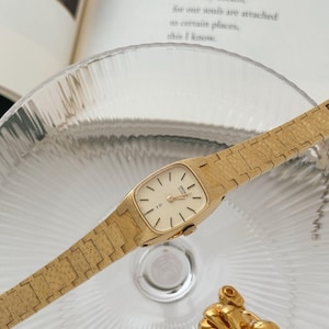 May include: A gold-coloured vintage watch with a rectangular face and a matching link band. The watch has a cream-coloured dial with black markers and hands. A gold-coloured octopus-shaped jewellery piece is also visible.