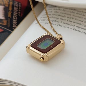Puede incluir: Un collar con colgante de reloj ALBA en tono dorado. La esfera rectangular del reloj tiene un borde rojo oscuro y una pantalla digital. El collar est&aacute; sobre un libro abierto con la palabra "HEMINGWAY" visible.