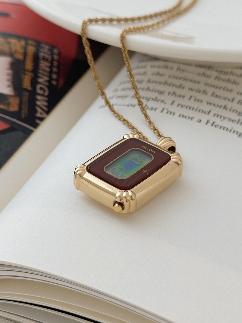 Puede incluir: Un collar con colgante de reloj ALBA en tono dorado. La esfera rectangular del reloj tiene un borde rojo oscuro y una pantalla digital. El collar est&aacute; sobre un libro abierto con la palabra "HEMINGWAY" visible.