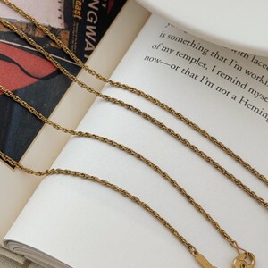 Puede incluir: Collar dorado con un dise&ntilde;o de cadena delicada. El collar se muestra en un libro abierto con el texto "Moveable Feast" visible. El cierre es visible en la parte inferior de la imagen.