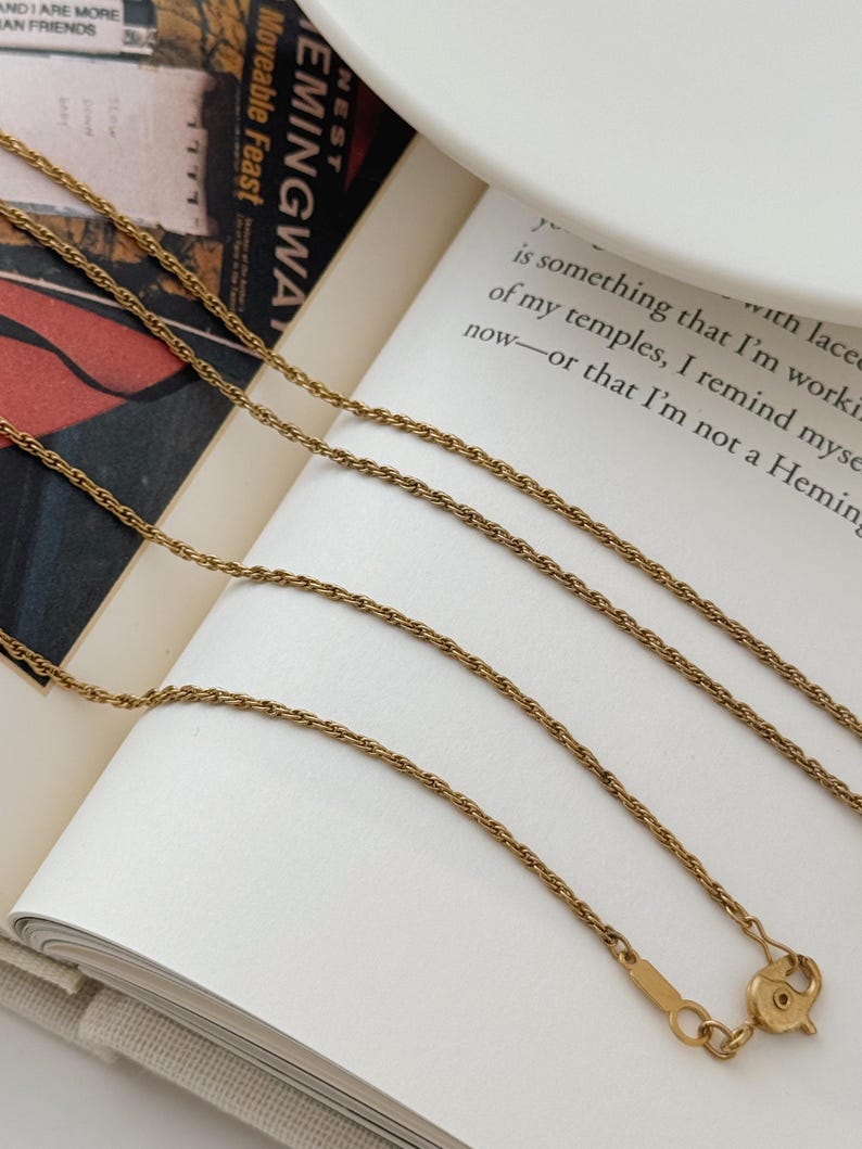 Puede incluir: Collar dorado con un dise&ntilde;o de cadena delicada. El collar se muestra en un libro abierto con el texto "Moveable Feast" visible. El cierre es visible en la parte inferior de la imagen.