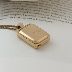 Puede incluir: Un collar con colgante rectangular dorado con una cadena delicada. El colgante tiene un peque&ntilde;o compartimento con bisagras y texto grabado. El collar se muestra en una p&aacute;gina de libro.