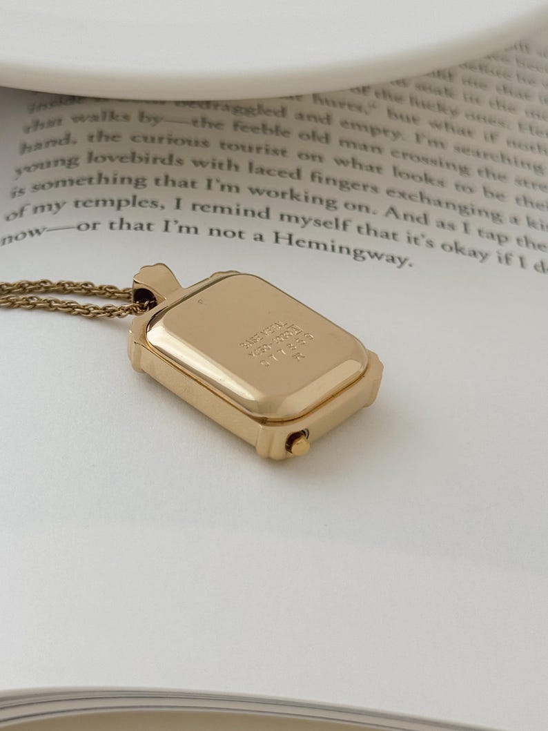 Puede incluir: Un collar con colgante rectangular dorado con una cadena delicada. El colgante tiene un peque&ntilde;o compartimento con bisagras y texto grabado. El collar se muestra en una p&aacute;gina de libro.