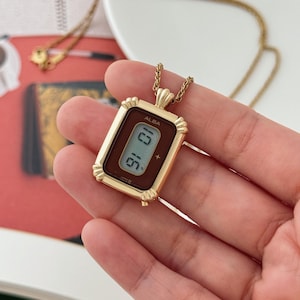 Puede incluir: Collar con colgante ALBA dorado con una esfera rectangular que muestra la hora digitalmente. La esfera del reloj tiene un fondo marr&oacute;n. El collar cuelga de una cadena dorada.