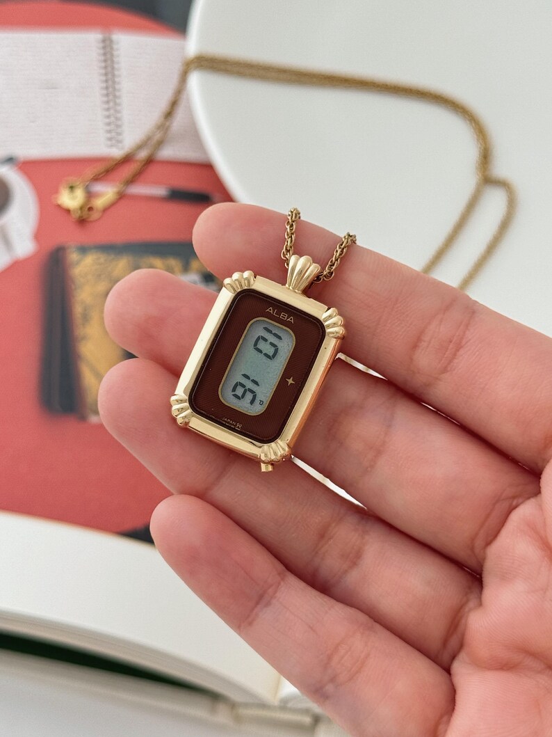 Puede incluir: Collar con colgante ALBA dorado con una esfera rectangular que muestra la hora digitalmente. La esfera del reloj tiene un fondo marr&oacute;n. El collar cuelga de una cadena dorada.