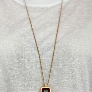 Puede incluir: Un collar dorado con un colgante rectangular que muestra la hora 11:56 en una pantalla digital. El colgante tiene una esfera burdeos y un borde dorado. El collar cuelga sobre una camiseta blanca.
