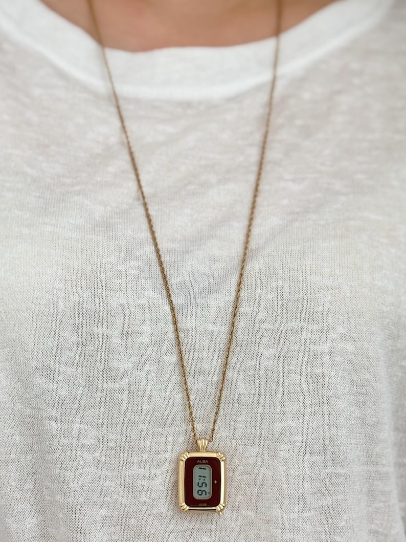 Puede incluir: Un collar dorado con un colgante rectangular que muestra la hora 11:56 en una pantalla digital. El colgante tiene una esfera burdeos y un borde dorado. El collar cuelga sobre una camiseta blanca.
