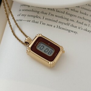 Puede incluir: Un collar con colgante rectangular dorado con una esfera de reloj digital. La esfera muestra la hora "10:08" en formato digital. La marca "ALBA" es visible. El collar est&aacute; sobre una superficie blanca.