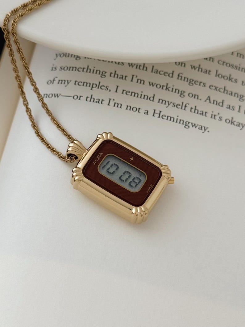 Puede incluir: Un collar con colgante rectangular dorado con una esfera de reloj digital. La esfera muestra la hora "10:08" en formato digital. La marca "ALBA" es visible. El collar est&aacute; sobre una superficie blanca.