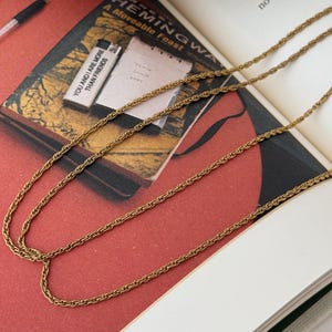 Puede incluir: Collar de cadena dorada con un delicado dise&ntilde;o de cuerda, sobre un libro abierto. La portada del libro muestra el t&iacute;tulo "A Moveable Feast" y el texto "YOU AND I ARE MORE THAN FRIENDS". El fondo es rojo y blanco.