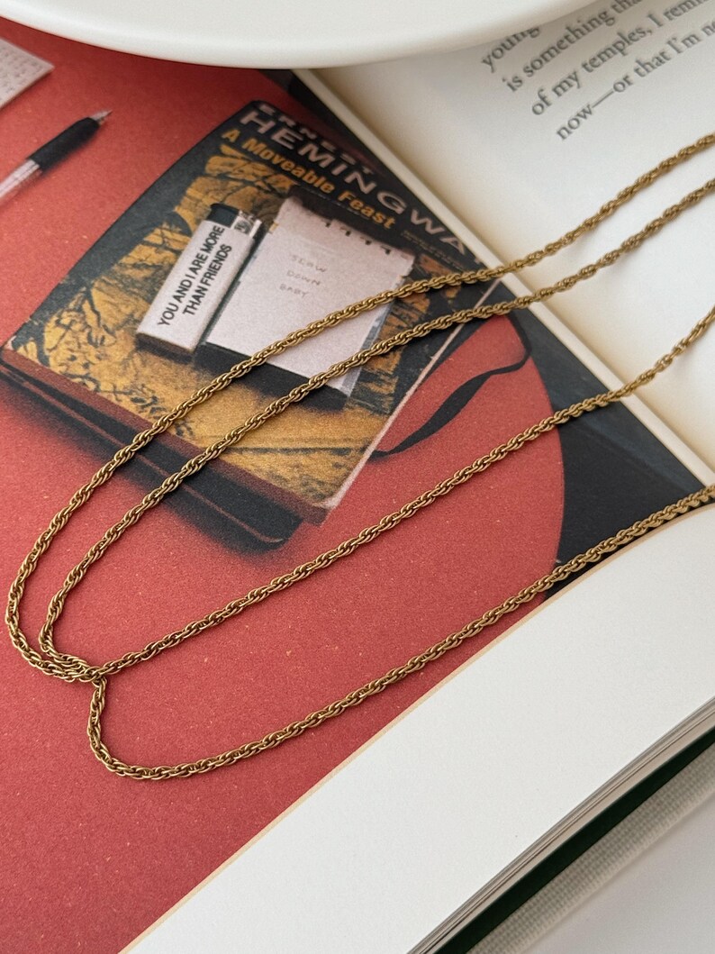 Puede incluir: Collar de cadena dorada con un delicado dise&ntilde;o de cuerda, sobre un libro abierto. La portada del libro muestra el t&iacute;tulo "A Moveable Feast" y el texto "YOU AND I ARE MORE THAN FRIENDS". El fondo es rojo y blanco.