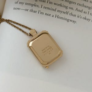 Puede incluir: Un collar con colgante rectangular de color dorado con una cadena delicada. El colgante tiene un marco texturizado y la parte posterior del colgante tiene el texto "BASE METAL Y450-4030 077859 R". El collar est&aacute; sobre una superficie blanca.