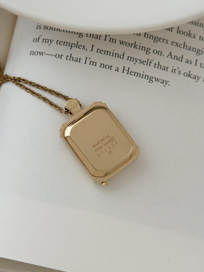 Puede incluir: Un collar con colgante rectangular de color dorado con una cadena delicada. El colgante tiene un marco texturizado y la parte posterior del colgante tiene el texto "BASE METAL Y450-4030 077859 R". El collar est&aacute; sobre una superficie blanca.