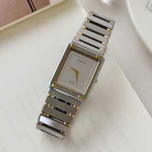 May include: A rectangular Lassale quartz watch with a silver-tone band and a gold-tone bezel. The watch face is light gray with gold hands and the word "Lassale" printed on the face. The watch is displayed on a white surface.