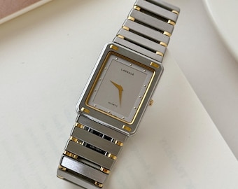 Könnte beinhalten: Rechteckige Lassale-Quarzuhr mit silberfarbenem Armband und goldfarbenen Details. Das Zifferblatt ist hellgrau mit goldenen Zeigern und dem Wort "Lassale". Die Uhr wird auf einer weißen Oberfläche präsentiert.