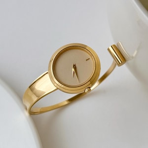 May include: A gold-toned bangle watch featuring a round case with a solid beige dial. The watch face has a single hour marker at the twelve o'clock position and two slender gold hands. The band is a polished open-cuff style with a cylindrical end.