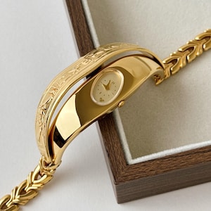 May include: A gold-tone wristwatch featuring an oval-shaped cream dial with gold hands and markers. The watch case is integrated into a wide, polished gold-tone cuff with an engraved pattern on the outer edge and a textured link bracelet strap.
