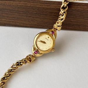May include: A gold-tone Seiko wristwatch featuring a round, textured gold dial with gold hands and a small dot marker. The watch case is accented with two small, round pink stones at the top and bottom, attached to a twisted gold-tone metal chain link band.