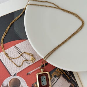 Puede incluir: Un collar dorado con un colgante rectangular que muestra "90:01". El colgante tiene una esfera burdeos y un marco decorativo. El collar est&aacute; sobre un plato blanco y una superficie roja. La marca "ALBA" es visible.