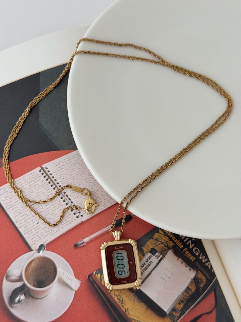 Puede incluir: Un collar dorado con un colgante rectangular que muestra "90:01". El colgante tiene una esfera burdeos y un marco decorativo. El collar est&aacute; sobre un plato blanco y una superficie roja. La marca "ALBA" es visible.