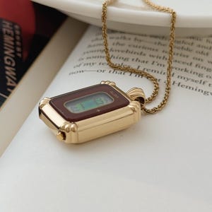 Puede incluir: Un collar con colgante dorado con una esfera de reloj rectangular con una pantalla burdeos. La esfera del reloj tiene la marca "ALBA" impresa. El collar est&aacute; sobre un libro abierto con texto.