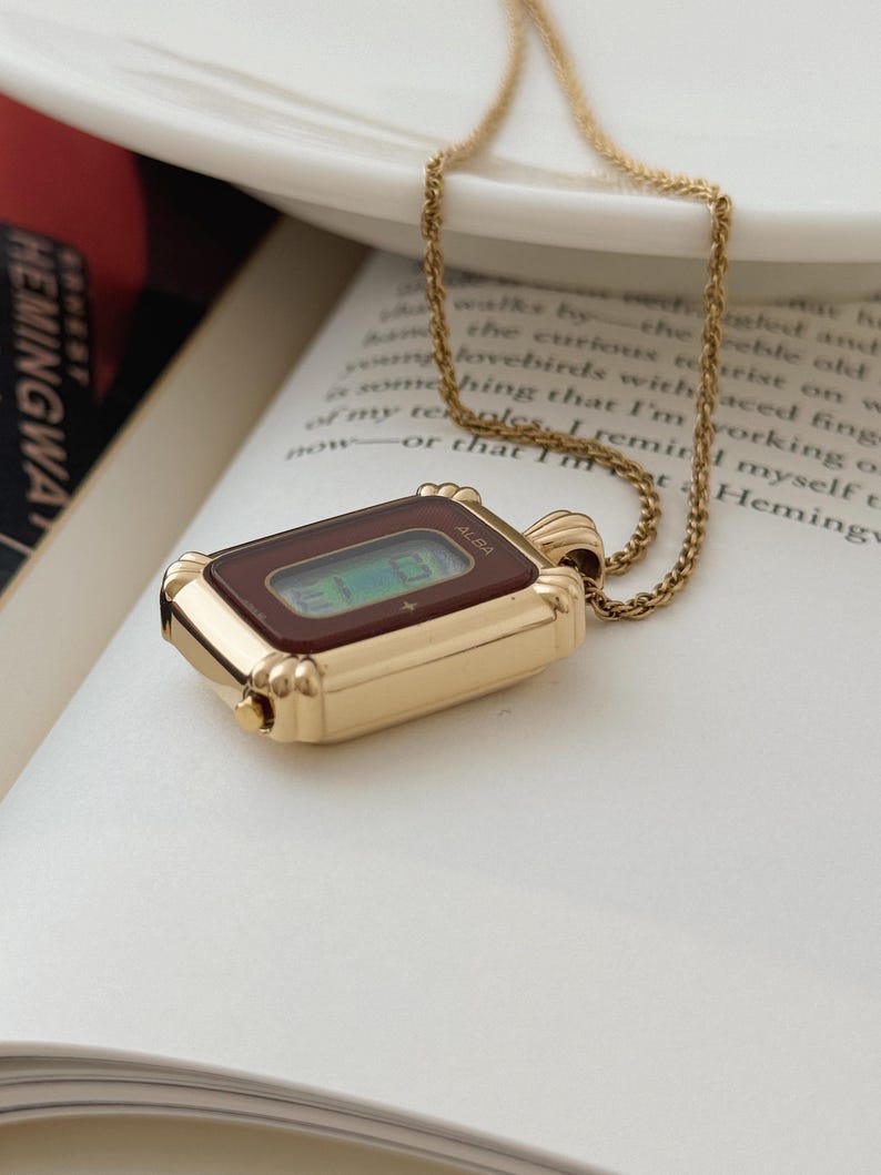 Puede incluir: Un collar con colgante dorado con una esfera de reloj rectangular con una pantalla burdeos. La esfera del reloj tiene la marca "ALBA" impresa. El collar est&aacute; sobre un libro abierto con texto.