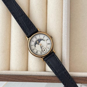 Peut inclure: Une montre dorée avec un bracelet en cuir noir. Le cadran de la montre est blanc avec des chiffres romains et un indicateur de phase lunaire. La montre a un petit cadran de secondes. L'ensemble est posé sur un fond neutre.