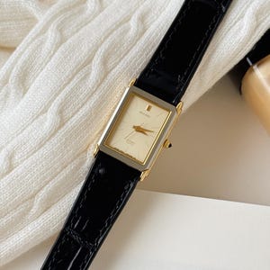 May include: A rectangular Citizen Exceed quartz wristwatch featuring a gold-tone dial with thin gold hands and hour markers. The watch has a two-tone silver and gold case and a black leather strap with a textured, embossed crocodile-skin pattern.