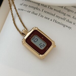 Puede incluir: Un collar dorado con un colgante rectangular que muestra la hora 10:12 en una esfera de reloj digital. La esfera del reloj es de color burdeos oscuro y el colgante est&aacute; inscrito con la marca "ALBA". El collar descansa sobre una superficie blanca.