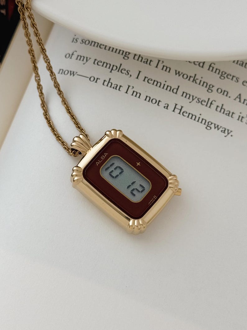 Puede incluir: Un collar dorado con un colgante rectangular que muestra la hora 10:12 en una esfera de reloj digital. La esfera del reloj es de color burdeos oscuro y el colgante est&aacute; inscrito con la marca "ALBA". El collar descansa sobre una superficie blanca.